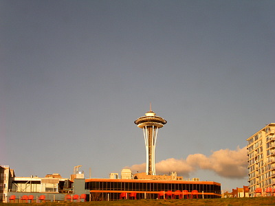 [A two story building with black windows and orange banding is in front of the tall structure of the Needle with the circular restaurant and other stuff at the top. The sky is blue with a few low-hanging white puffy clouds.]